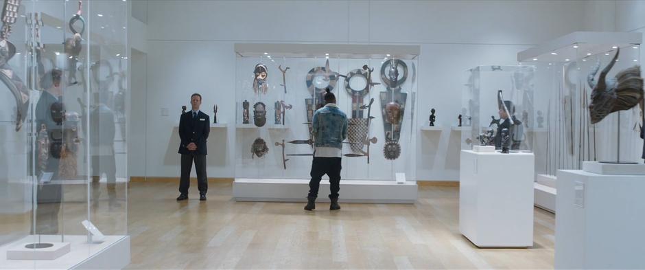 Killmonger stands in front of a display case of African artifacts while several security guards keep an eye on him.