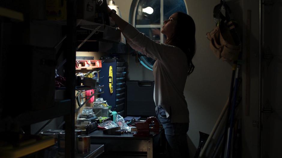 Sam reaches to a high shelf to grab a crowbar.