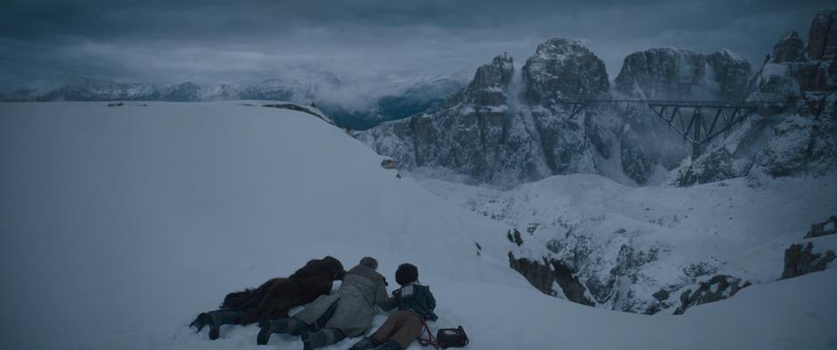 Han, Beckett, and Val lie down on the edge of the cliff and examine the bridge in the distance.