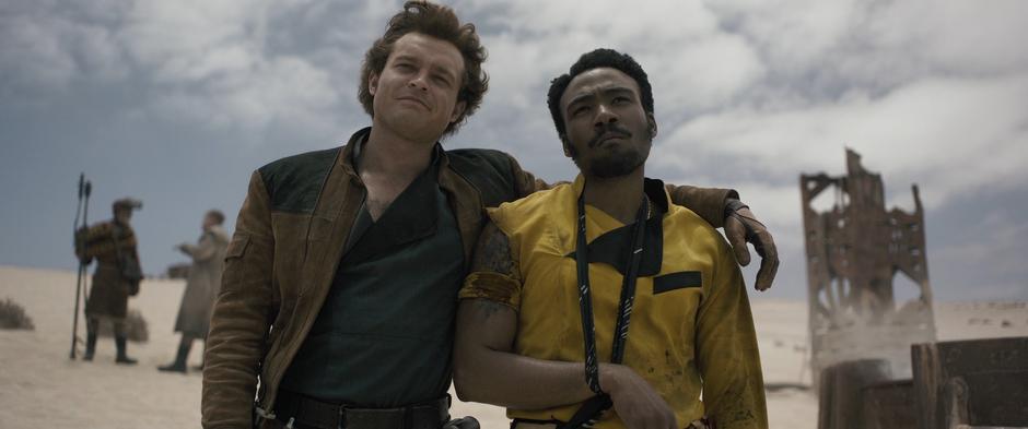 Han puts his arm around Lando's shoulder as they both look down at the Falcon.