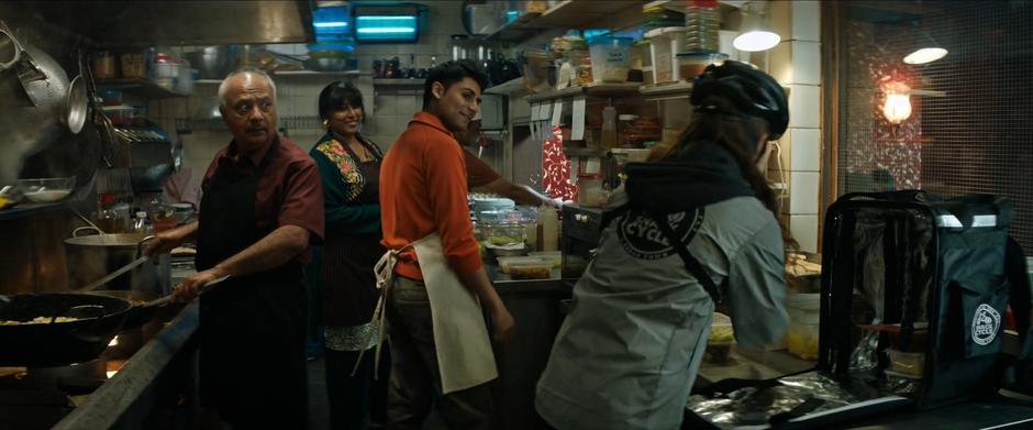 The three people in the kitchen look over at Lara as she packs up the food in her backpack.