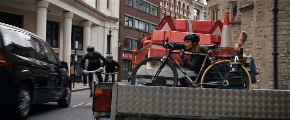 Lara watches as several bicyclists pass as she hides on the back of the trailer.