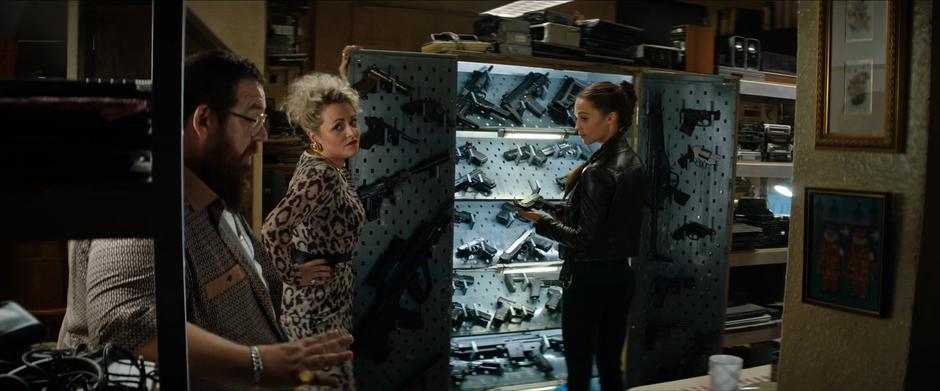 Lara examines a pistol from the weapon case in the back of the pawn shop while Alan and his wife talk.