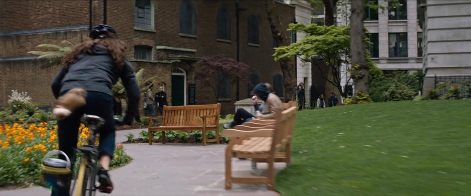 Lara bikes through a narrow path past some benches.