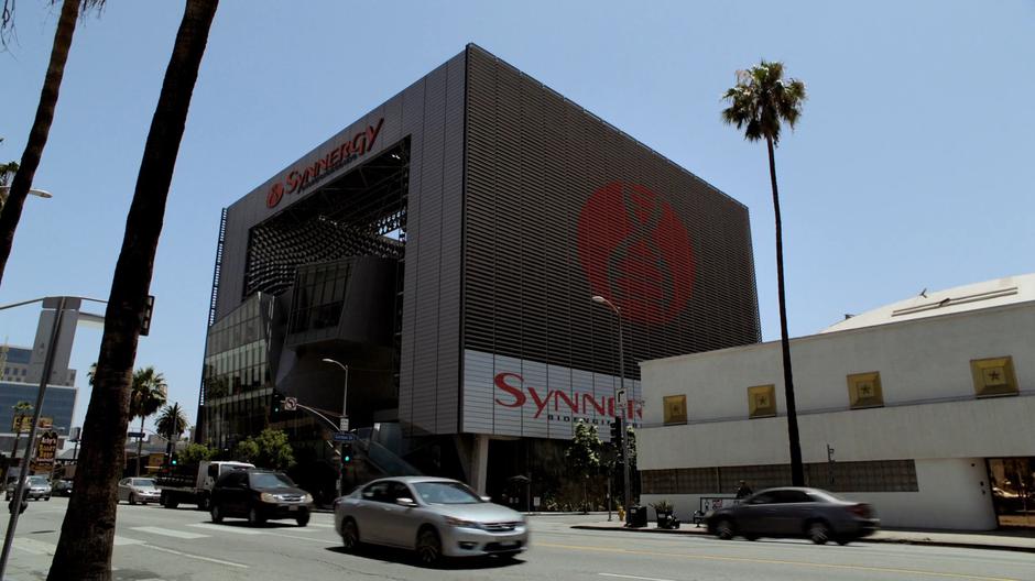 Establishing shot of the Synnergy building as cars drive past on the street.