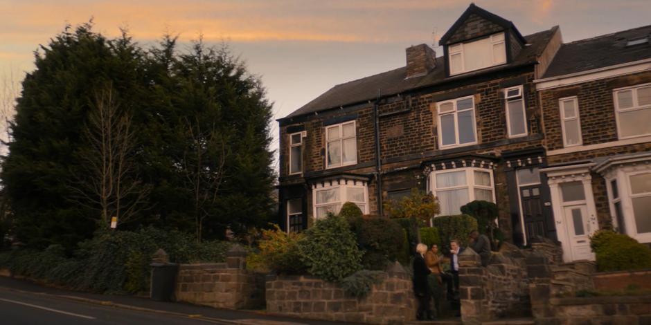 The Doctor, Yasmin, Graham, and Ryan sit on the steps in front of the flat talking and drinking tea.