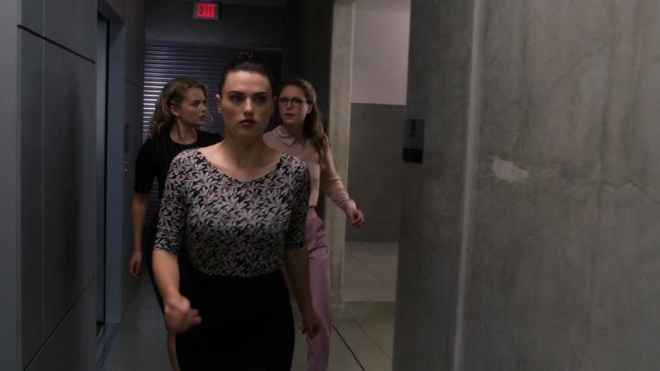 Lena leads Eve and Kara down the hallway away from the elevator.