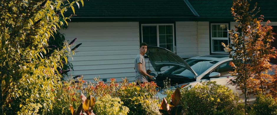 John looks over at Lara Jean's house while he works on the engine of his car.