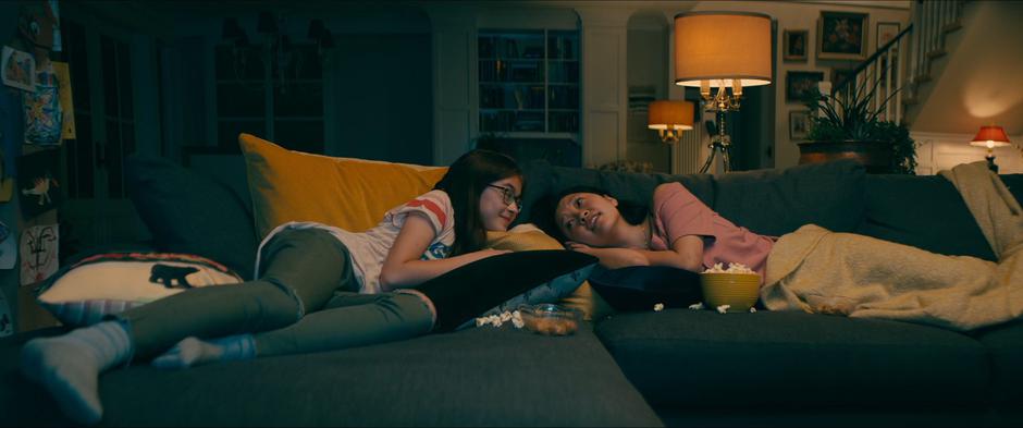 Kitty and Lara Jean talk while lying on the couch watching a movie.