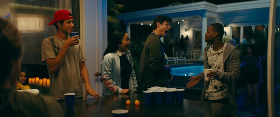 Lara Jean stands with Peter as he talks to Greg who is playing beer pong.