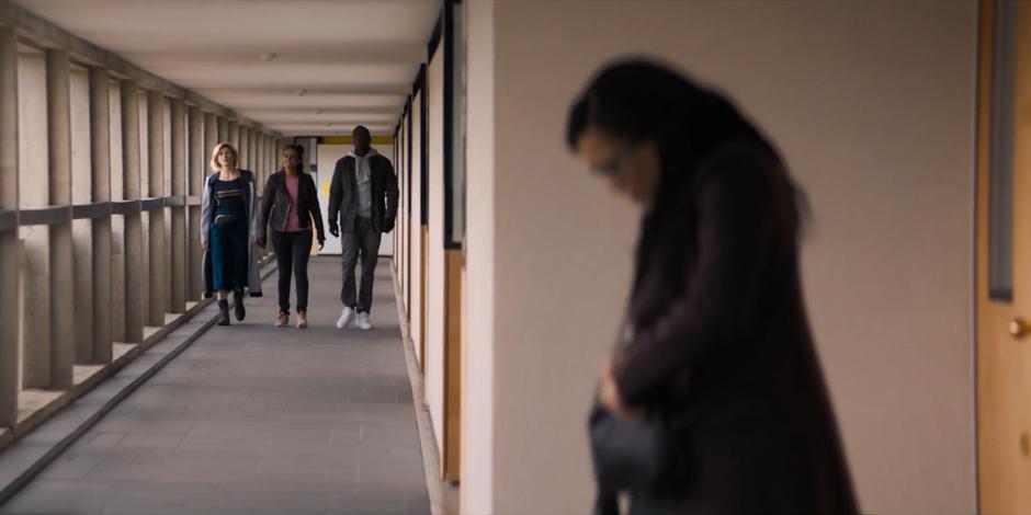 The Doctor, Yasmin, and Ryan walk down the hallway as Jade stands outside the neighboring flat.