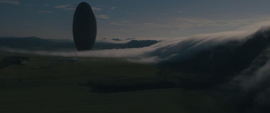 The alien craft hovers over the field as fog rolls in over the hills.