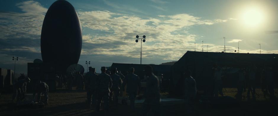 Members of the military move around the camp in the foreground while the alien craft hovers behind them.