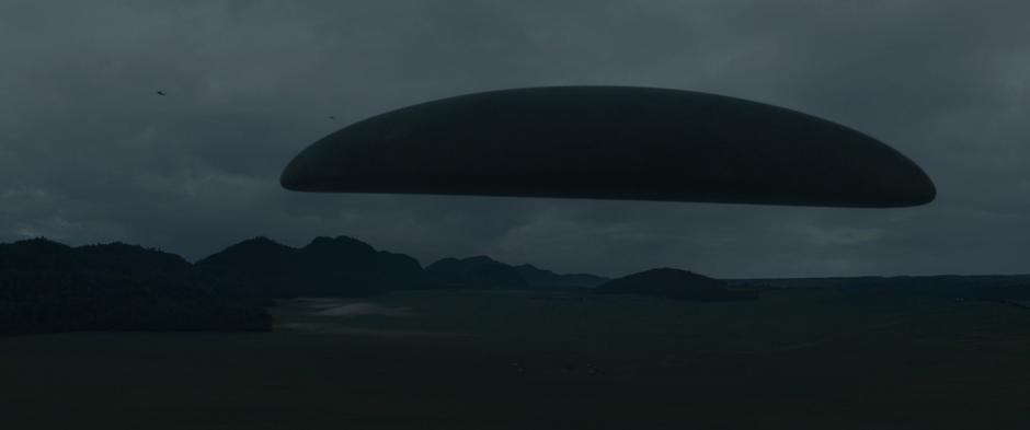 The alien craft turns horizontal over the landscape as several helicopters hover overhead.