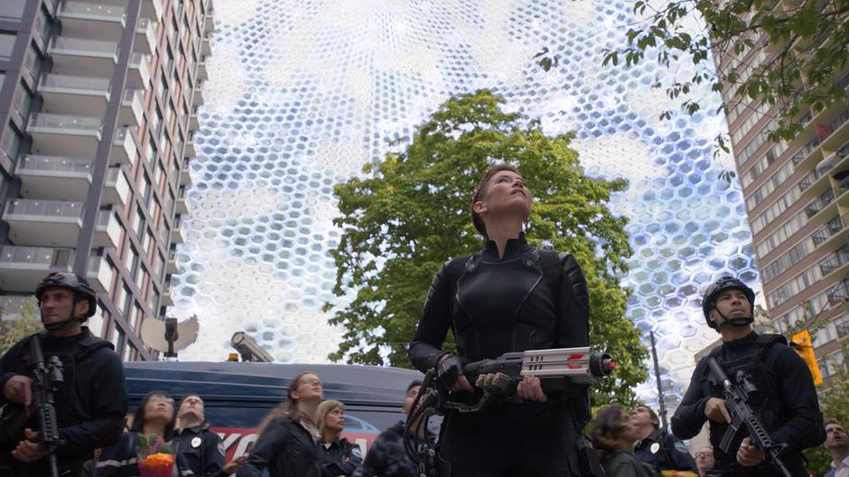 Alex looks up at the bubble forming over the block while standing with two DEO agents.