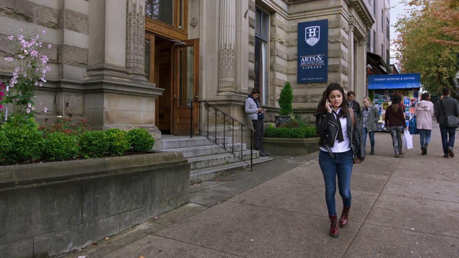 Mel talks to Niko on the phone while walking in front of the university library.