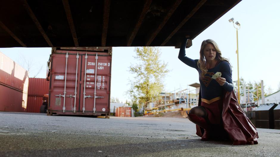 Kara lifts up the shipping container with one arm while examining the shipping label she found underneath it.