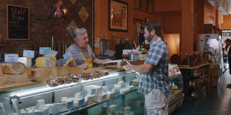 The store employee hands Frank a job application from behind the cheese counter.