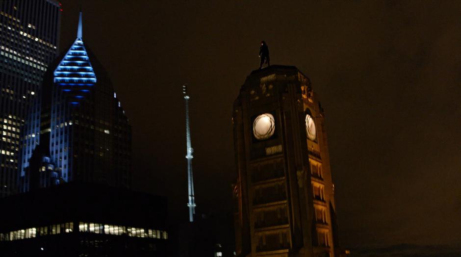 Batwoman stands on top of the Wayne Enterprises spire looking down on the lower rooftop.