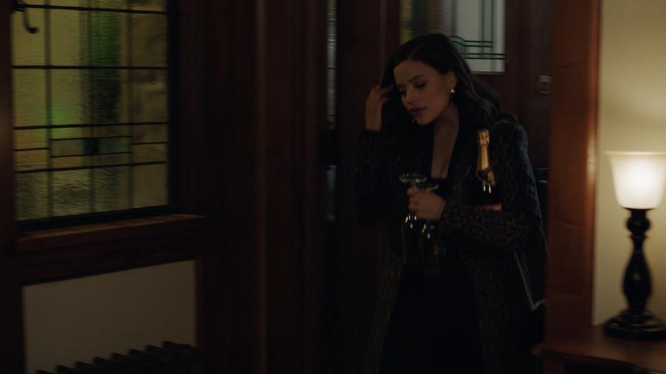 Maggie walks into the back hallway holding a bottle of wine with two glasses.