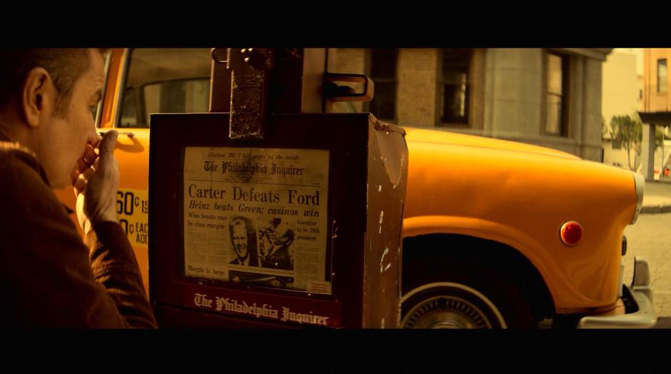 Victor kneels down at the newspaper dispenser which contains a paper from '76.