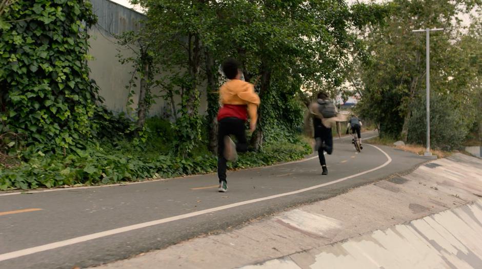 Alex and Chase follow after Mike on a Bike as he rides up the side path towards the street.