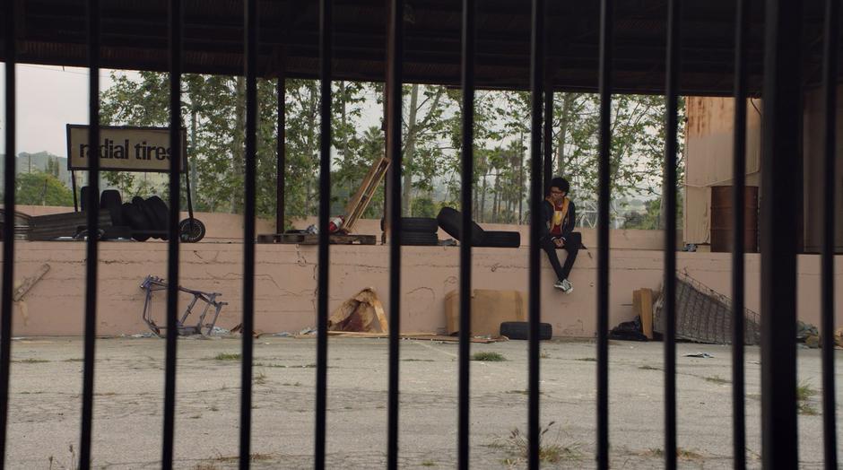 Alex glances over from where he is sitting on the abandoned loading dock as Darius's car approaches.