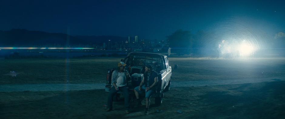 Two guys sit on the back of a pickup truck as Cable's pod crashes into the field behind them.