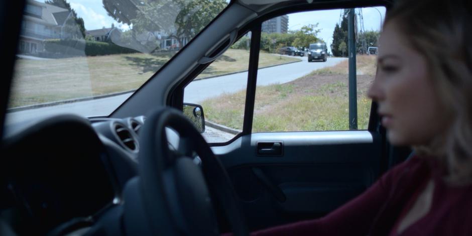 Marcy waits in her vehicle while the police van drives down the street towards her.