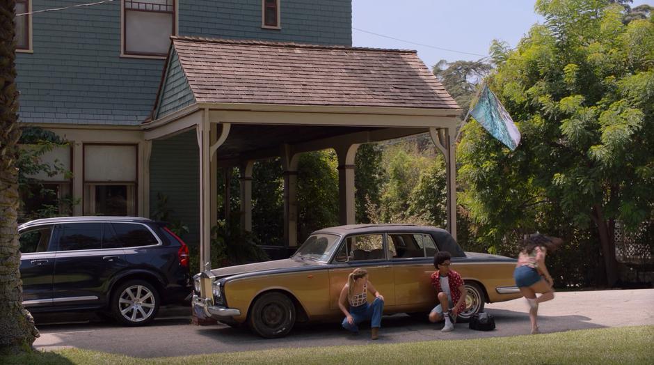 Karolina and Alex kneel behind the car while Molly runs around towards the house.