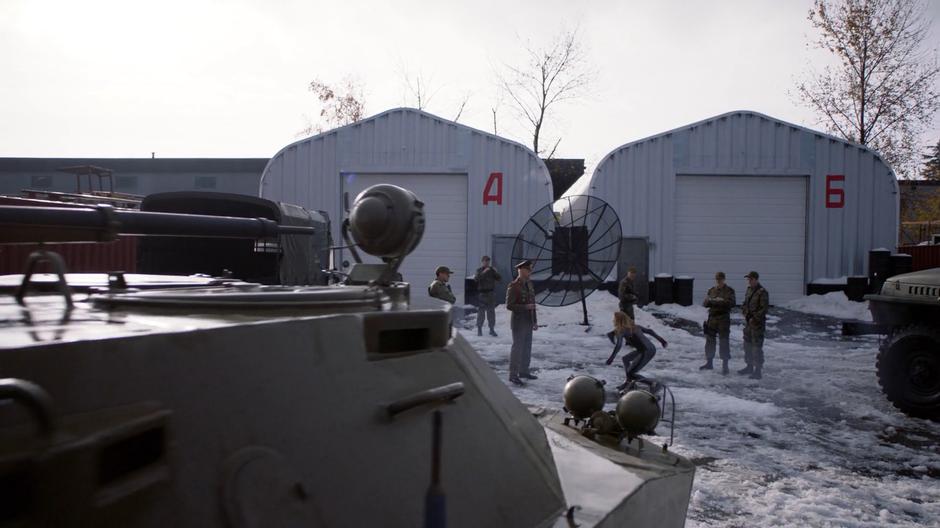 The Kasnian Supergirl lands in front of her handler in a snowy lot filled with other soldiers.