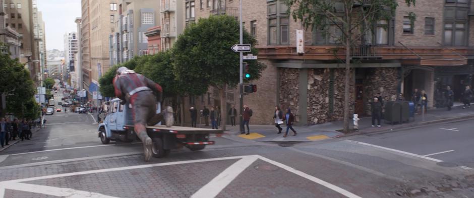 The enlarged Scott kneels on the back of the pickup truck and rides it down the hill.