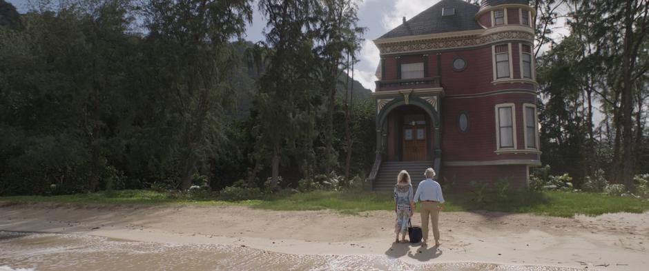 Hank reaches for Janet's hand as they stand in front of their house sitting on the edge of the beach.