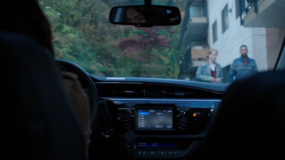 Nicole watches people walk past her car as she talks to her boss on the phone.