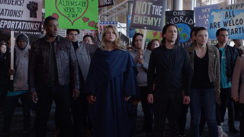 J'onn, Kara, Brainy, and Nia stand at the front of the protestors.