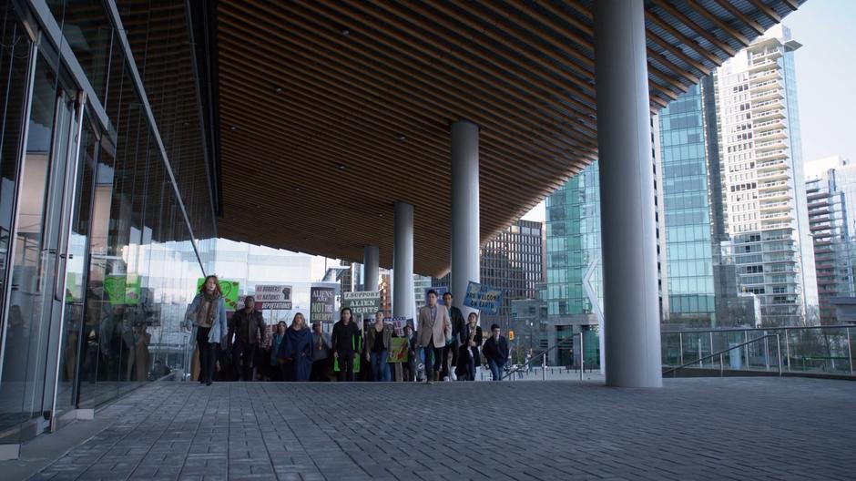 J'onn, Kara, Brainy, and Nia walk at the front of the alien march up the steps to the door of the Department of Alien Affairs.