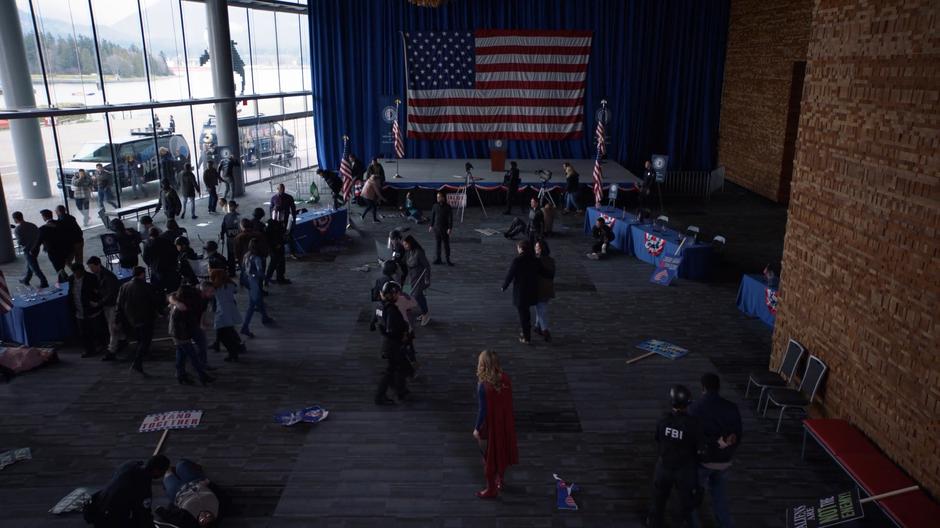 Kara stands in the middle of the room as the DEO helps clean up fight.