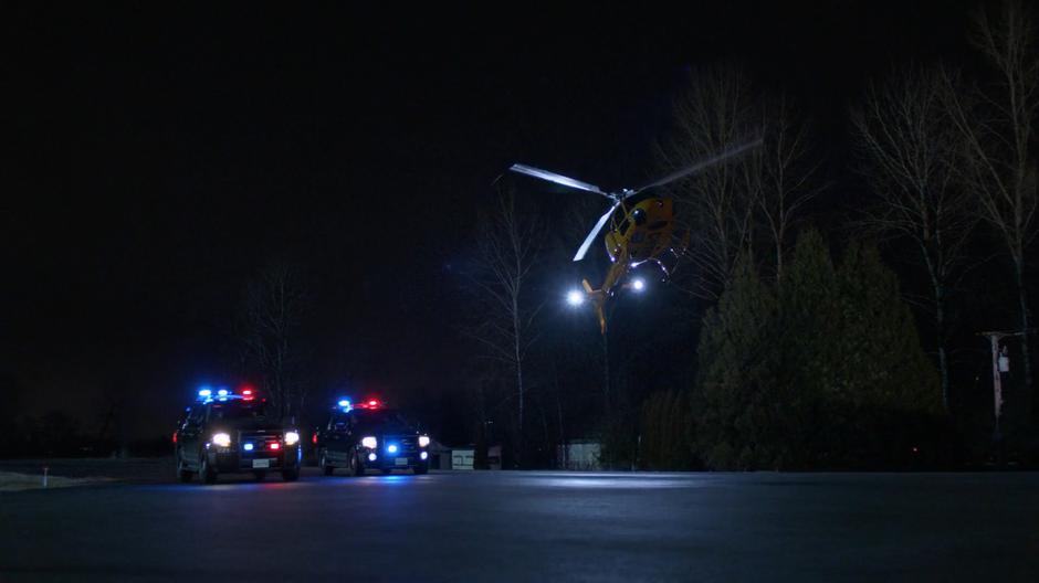 Two police cars pull up with lights flashing as the medivac helicopter lands on the pavement at night.