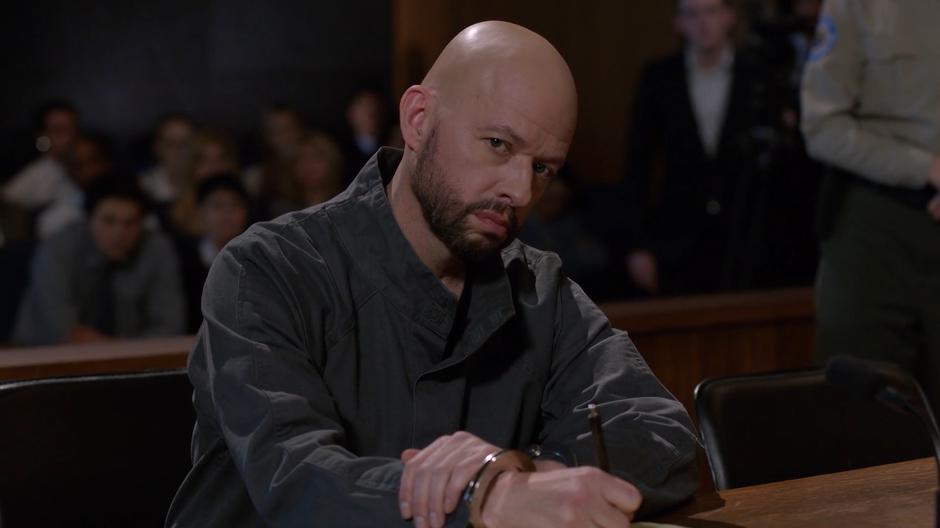 Lex looks up at Lena during her testimony while writing on a notepad.