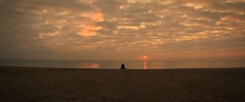 Sarek kneels on the beach meditating while the sun rises over the water.