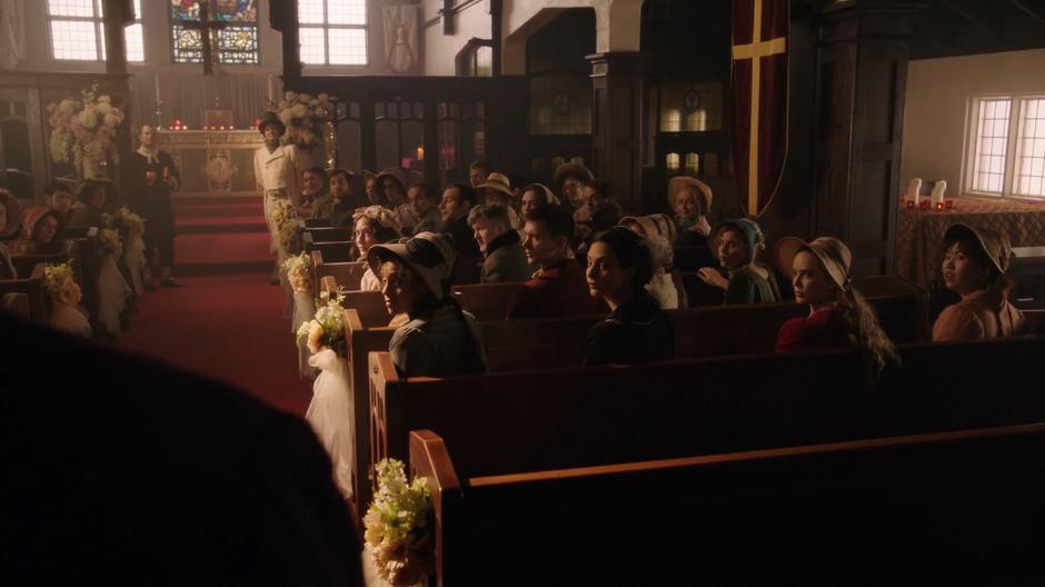 Charlie, Zari, Sara, Jane Austen, Mona, and the other attendees turn around to watch as the bride enters on her father's arm.