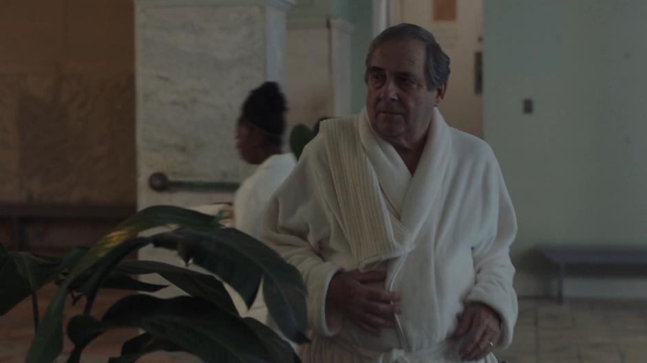 Senator Henderson walks towards the steam room wearing a robe.