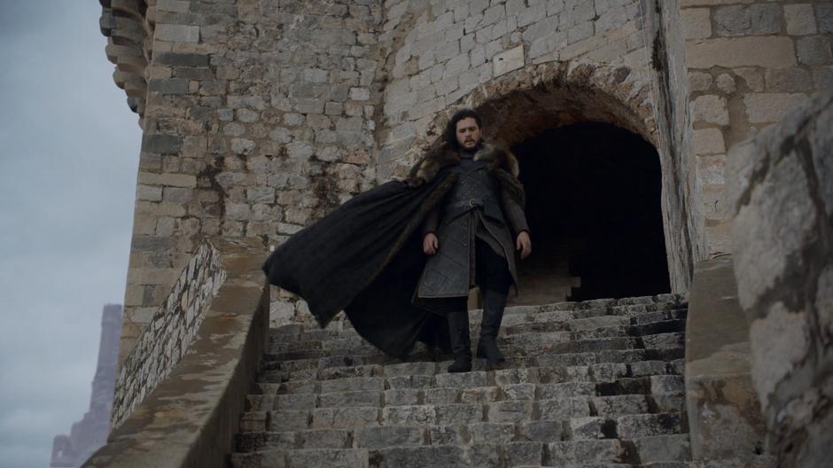 Jon walks down the steps from the castle with his cloak blowing in the wind.