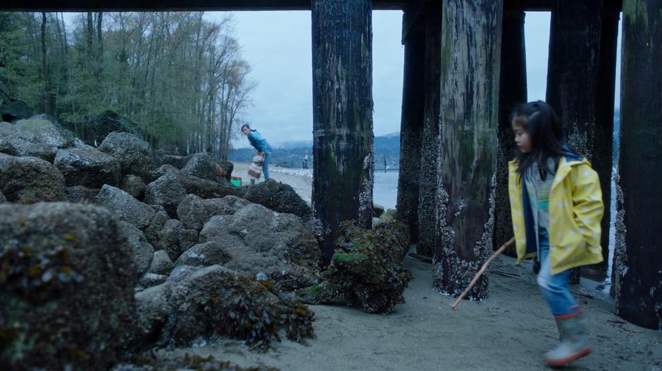 A young girl plays with a stick on the beach while her mother keeps an eye on her while packing up their stuff in the distance.