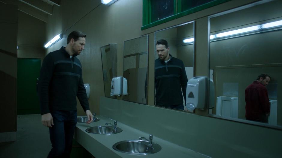 Rick glances over his shoulder from the urinal while a mysterious man approaches the sinks.