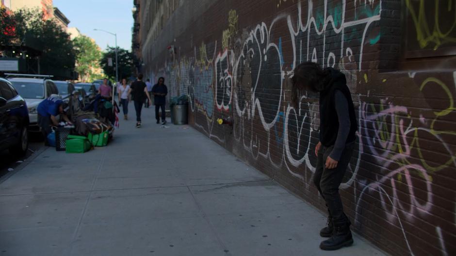 A drunk guy leans against the graffiti-covered wall as Trish walks past.