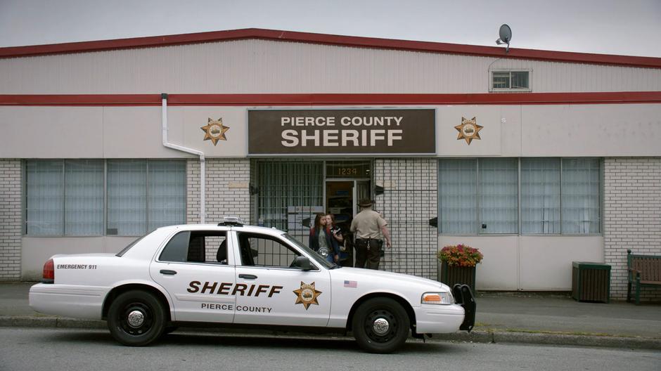 The deputy closes the door of the station while leading Pippi and Annie out to his squad car.