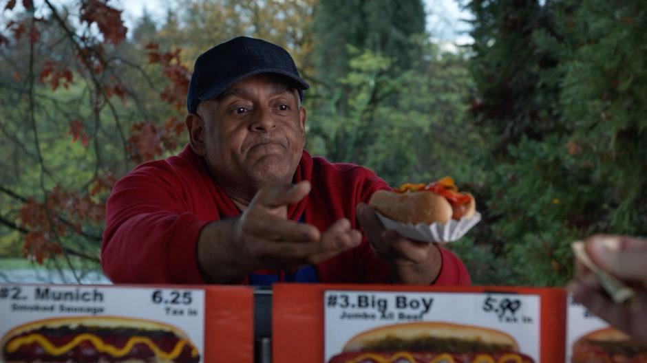 The hot dog vendor reaches for the cash and hands over one of the hot dogs.