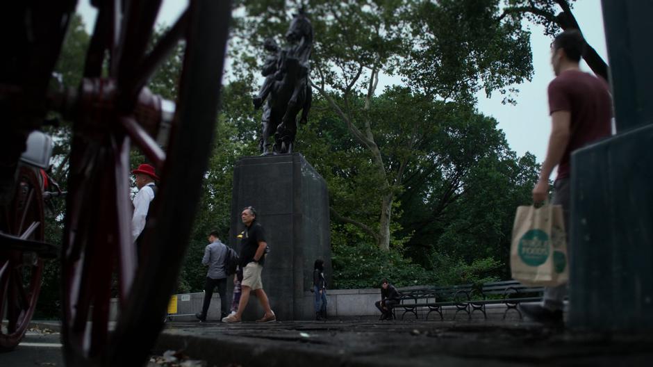 Jessica stands by the statue while Erik reaches under the bench searching for the third drop.