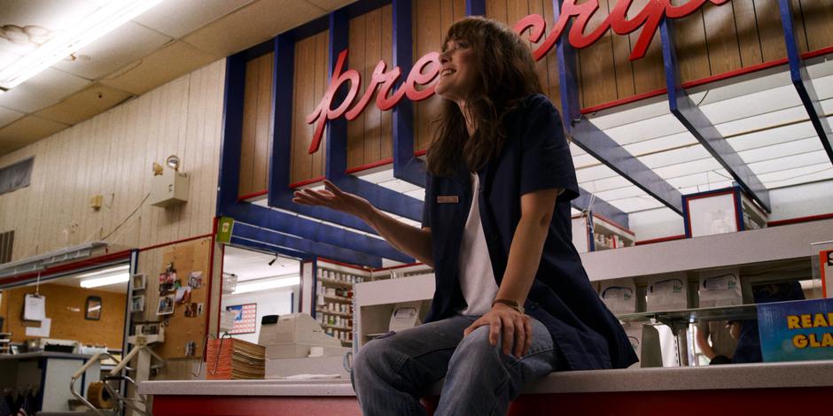 Joyce sits on the pharmacy desk counter while talking to Hopper.